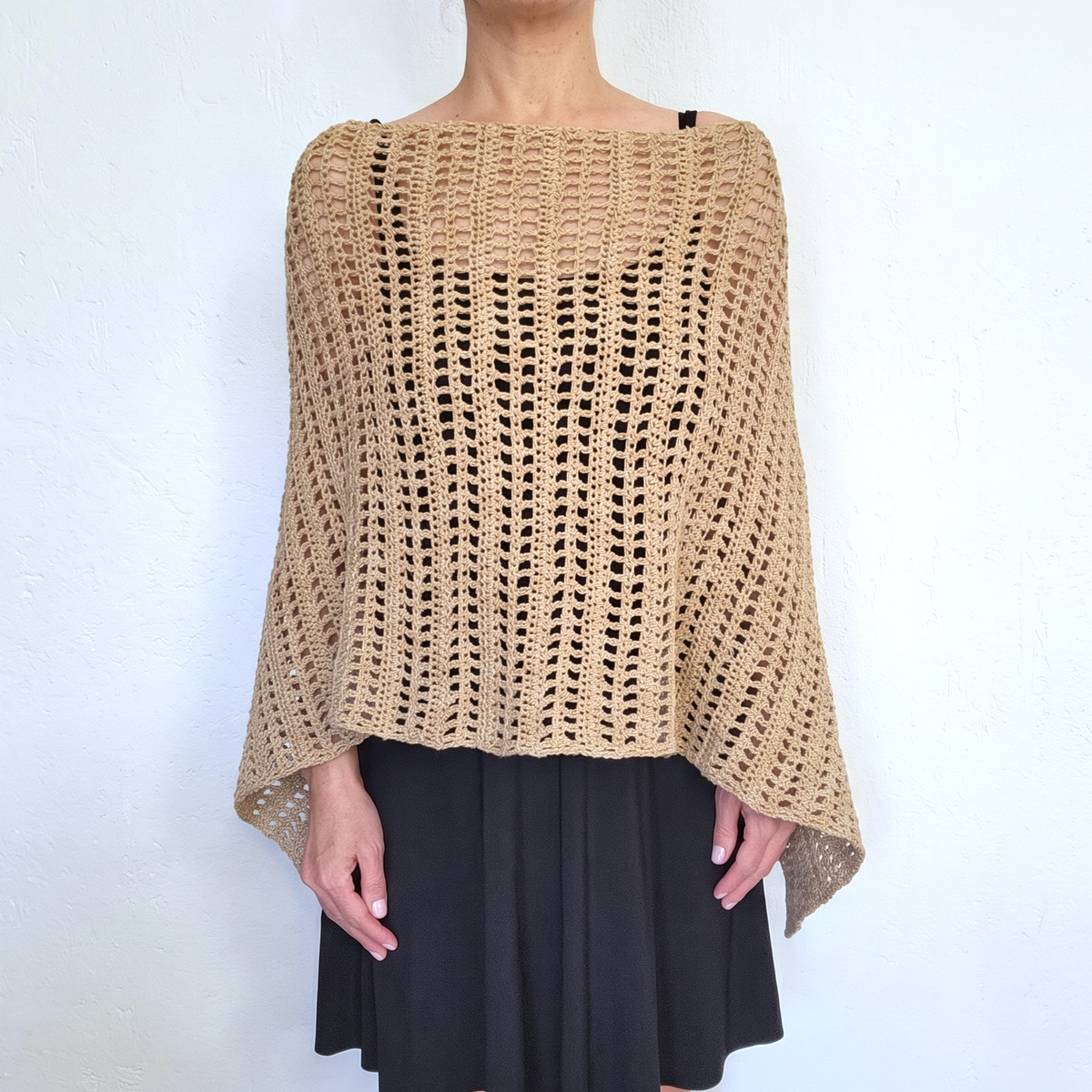 A person wearing a chunky open-knit beige crochet poncho over a black skirt, standing against a plain white wall.